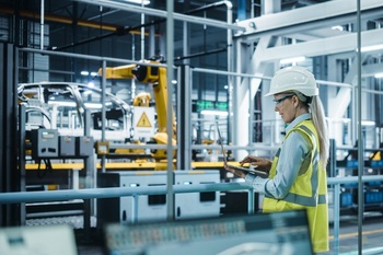 Car Factory: Female Automotive Engineer Wearing Hard Hat, Standing, Using Laptop. Monitoring, Control, Equipment Production. Automated Robot Arm Assembly Line Manufacturing Electric Vehicles.